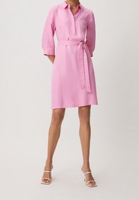 Abito camicia rosa con colletto, maniche tre quarti e cintura annodata. Tessuto liscio con orlo fino al ginocchio e dettagli minimi.