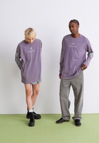 Deux personnes se tiennent devant un mur blanc, vêtues de t-shirts à manches longues violets assortis avec des logos, l'une en bottes et en shorts, l'autre en pantalon ample et en baskets.