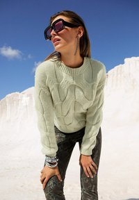 Frau mit großen schwarzen Sonnenbrillen, langen hängenden Ohrringen, einem blassgrünen, grobstrickigen Pullover und Schlangenhautmuster-Hosen vor einem strahlend blauen Himmel.