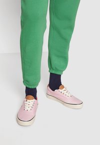 Světle růžové plátěné tenisky s bílými tkaničkami a krémovými podrážkami, obuté s námořnickými ponožkami a zelenými zúženými joggingovými kalhotami s elastickými manžetami.