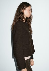 Brown textured jacket featuring a round neckline, front button closure, and two pockets. Styled with a light pink top underneath.