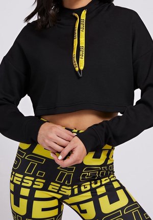 Kobieta ubrana w czarną krótka bluzę z kapturem z żółtymi sznurkami oraz czarne legginsy z wysokim stanem ozdobione wyraźnym żółtym napisem graficznym.