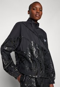 Black zip-up jacket featuring a textured, reflective snake-skin design. Smooth fabric on upper panels, elastic cuffs, and a stand collar.