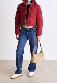 Kobieta w skróconej, czerwonej kurtce puchowej, w czarno-białym, paskowanym topie, niebieskich dżinsach i białych trampkach, trzymająca beżową torebkę.