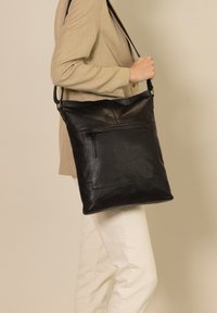 Borsa a spalla in pelle nera con texture liscia, tasca frontale con zip, forma piatta e tracolla regolabile, portata da una persona.