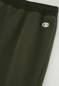 Dunkelgrüne Sweatpants mit einem elastischen Bund, strukturiertem Stoff und einem kleinen bestickten Logo am unteren linken Oberschenkel.