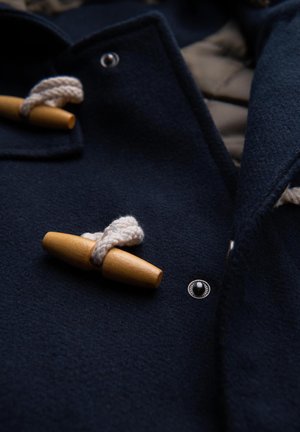 Cappotto di lana blu scuro con bottone a toggle in legno fissato attraverso un spesso anello di corda bianca e un bottone a pressione in metallo visibile.