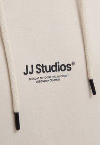Hellbeige Kapuzenpullover aus strukturiertem Stoff, mit einem schwarzen, gestickten Logo "JJ Studios" und grauen Kordeln mit schwarzen Enden.