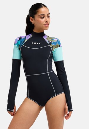 Vrouw die een zwarte wetsuit met lange mouwen draagt met turquoise en bloemmotief op de schouders, staand tegen een effen achtergrond.