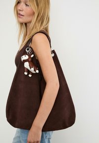 Donna bionda che indossa una canotta marrone senza maniche, porta una grande borsa a spalla in suede marrone con un piccolo portachiavi a forma di renna marrone e bianca attaccato.