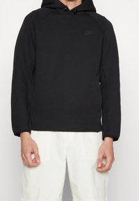 Svart hoodie med en strukturerad design, som har en framlogotyp, dragsnören för huvan och ribbstickade muddar och fåll. Mjuk tygtextur.