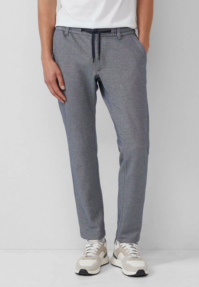 Homme portant un pantalon de jogging gris ajusté avec un cordon noir et des baskets blanches, debout sur un fond blanc.