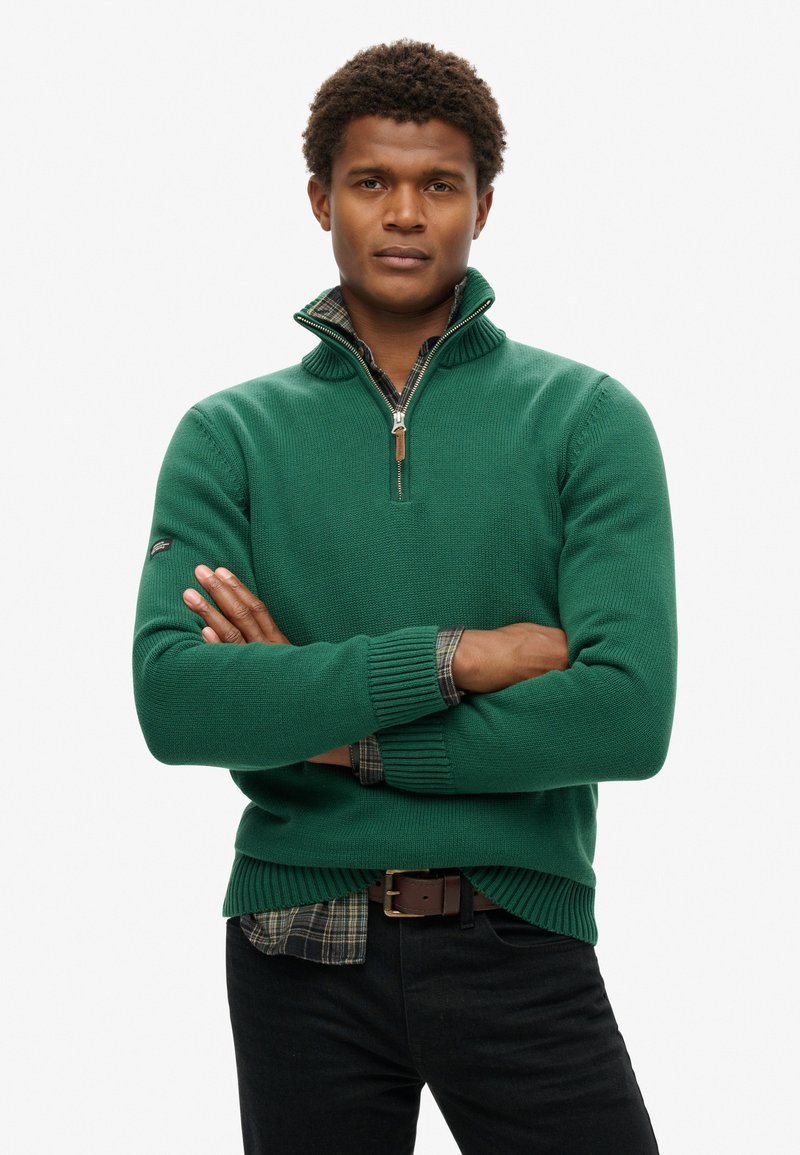 Grüner Strickpullover mit Zip-Kragen, gerippten Bündchen und Saum, getragen über einem karierten Hemd. Dunkle Jeans runden das Outfit ab.