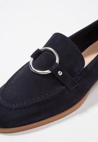 Mocassino in suedè blu navy con un accento metallico rotondo, dettagli in cucitura e suola in gomma marrone. La vista ravvicinata mette in evidenza la texture e il design.