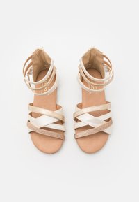 Sandaler med remmer og flat såle, med hvite, gullfargede, og glitrende bronsefargede bånd som krysser over foten, og en bakre ankelrem.