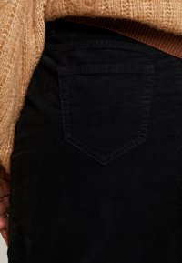 Zwarte corduroy broek met een enkele achterzak met stiksel, gedeeltelijk bedekt door een lichtbruin gestructureerd sweater.