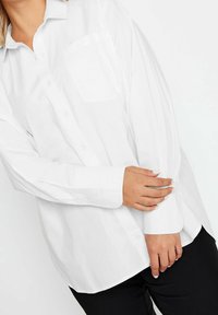 Woman wearing a loose white button-up shirt and black pants adjusting the cuff on her left sleeve with her right hand.