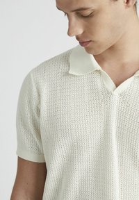 Polo en tricot color écru avec un motif texturé, doté d'un col doux et de manches courtes, présentant une coupe décontractée et un matériau léger.