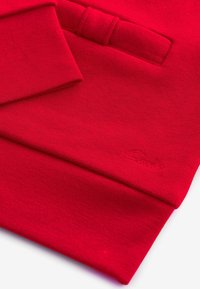 Rotes Sweatshirt mit glatter Textur, das ein kleines gesticktes Logo und ein umgeschlagenes Bündchen-Design aufweist. Hergestellt aus weichem, gestricktem Material.