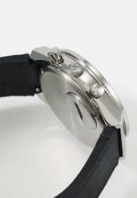 Montre en métal argenté avec un bracelet en cuir noir, présentant une lunette texturée, des boutons-pousseurs et une forme de boîtier ronde et simple.