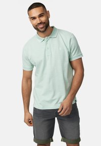 Lys grøn polo shirt med en subtil struktur, der har en hvid krave og en tre-knap lukning. Båret med mørkegrå shorts, der er rullet op ved ærmerne.