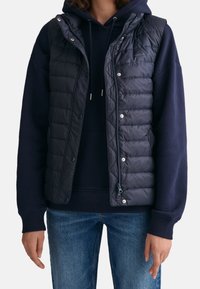 Marinblå quiltad pufferväst med huva, tryckknappar och dragkedja över en mörk sweatshirt, i kombination med ljusblå jeans.