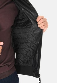 Giacca di pelle nera con interno in nylon trapuntato. Presenta una zip argentata, polsini a costine e una superficie esterna liscia e testurizzata.