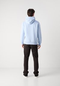 Homme debout de dos, portant un sweat à capuche bleu clair, un pantalon foncé et des baskets noires avec des semelles blanches sur un fond blanc uni.