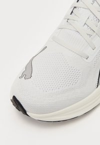 Zapatilla de correr blanca con parte superior de malla transpirable, patrón texturizado y suela de goma negra. Presenta una silueta de gato en el lateral.