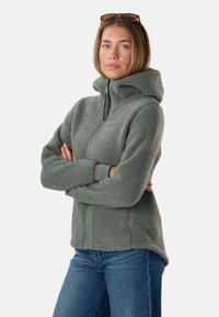 Frau mit verschränkten Armen trägt eine graue Fleecejacke mit Reißverschluss und Kapuze, blaue Jeans und eine Sonnenbrille auf dem Kopf, schaut nach vorne.