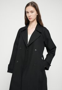 Melns tranšejmētelis ar dubultpogu dizainu, lielu apkakli, sānu kabatām un gludu tekstūru. Pielāgota piegrieztne ar pogu aizdari.