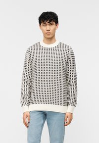 Jeune homme portant un pull en tricot crème et noir à motifs, avec des poignets et un ourlet côtelés, associé à un jean bleu clair, debout devant un fond blanc.