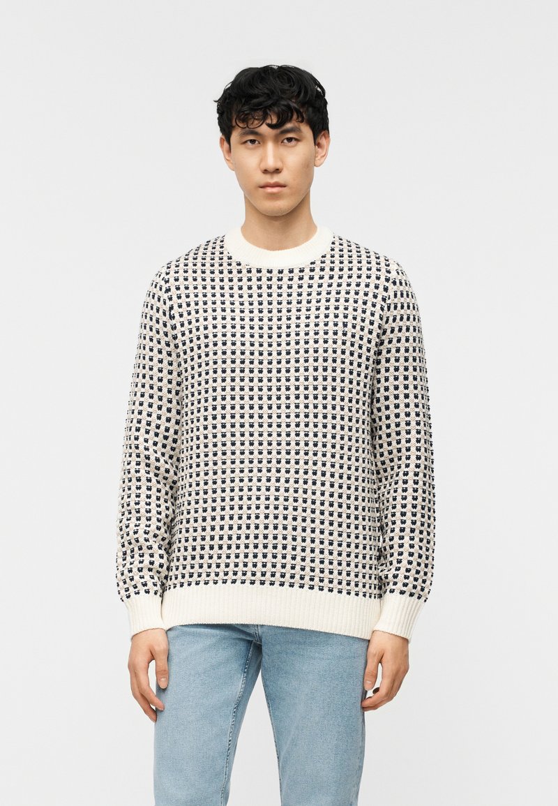 Jeune homme portant un pull en tricot crème et noir à motifs, avec des poignets et un ourlet côtelés, associé à un jean bleu clair, debout devant un fond blanc.