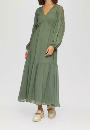 Vrouw die een lange olijfgroene jurk met V-hals draagt, met doorschijnende pofmouwen en beige chunky loafers met gouden kettingdetail.