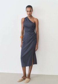 Rochie albastră cu un umăr, cu plisare laterală și fante adânci, realizată dintr-un material ușor. Textură netedă, până la șold, asortată cu pantofi slip-on bej.