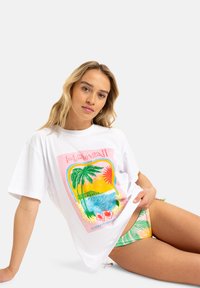 Hvit bomulls t-skjorte med et fargerikt hawaiiansk motiv som viser palmer, hav og sol. Løs passform, korte ermer og rund hals.