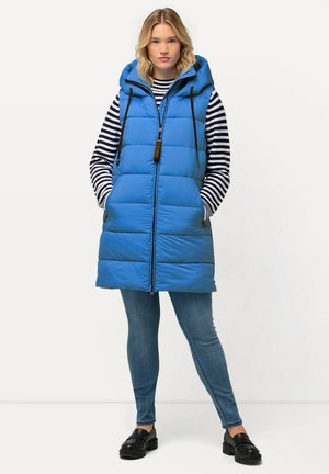 Blauer Steppweste mit Kapuze, ausgestattet mit einem Frontreißverschluss, zwei Seitentaschen und schwarzen Kordelzügen. Über einem gestreiften Langarmshirt und Jeans getragen.
