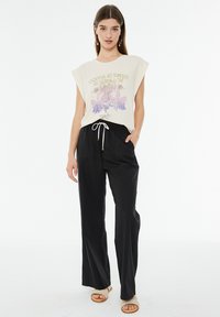 Camiseta gráfica beige con mangas tipo cap, estampado en púrpura y dorado. Pantalones anchos negros con cordón y sandalias beige claro.