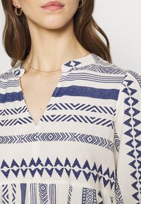 Vit klänning med marinblå geometriska mönster, med V-ringning och korta ärmar. Mjukt tyg med en något strukturerad yta.