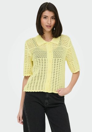 Mladá žena s rovnými tmavými vlasmi, oblečená v svetložltom háčkovanom krátkom rukáve s gombíkmi a čiernych vysokých nohaviciach.