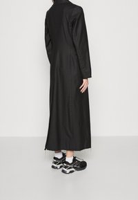 Long manteau noir en tissu lisse, doté d'un col et d'un détail de couture. Porté avec des baskets noires et des chaussettes blanches.