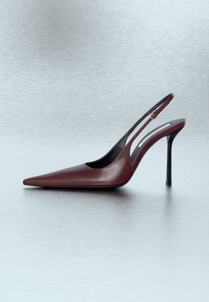 Décolleté slingback in pelle rossa scura con punta a punta, tacco alto sottile e cinturino regolabile, su superficie grigia testurizzata.