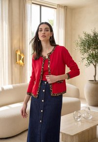 Nainen sisätiloissa, pukeutunut punaiseen neulejakkuun, jossa leopardikuvioinen reunus, yhdistettynä samansävyiseen toppiin ja tummansiniseen napitettuun farkkuhameeseen, seisoo beigen sohvan ja puisen pöydän lähellä.