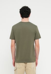 Βαμβακερό t-shirt σε ελιά πράσινο χρώμα με κοντά μανίκια, στρογγυλή λαιμόκοψη και απαλή υφή, εμφανιζόμενο από πίσω, συνδυασμένο με χακί παντελόνια.