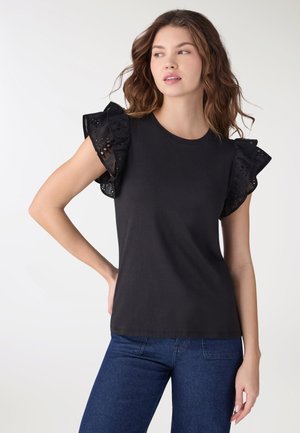 Donna che indossa una canotta nera senza maniche con maniche in pizzo a occhielli arricciate e jeans blu, in piedi con una mano sul fianco.