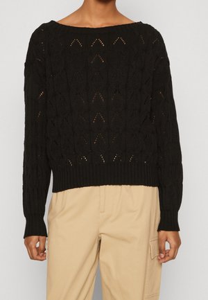 Persoon dragen een zwart gebreide trui met een chevronpatroon en beige cargobroek, staand tegen een effen achtergrond.