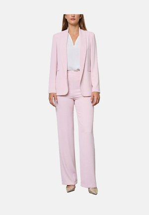 Vrouw die een lichtroze blazer en bijpassende wijde broek draagt, met een witte blouse en beige puntige schoenen, staand tegen een witte achtergrond.