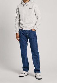 Persona con felpa grigia chiara con il logo "Pepe Jeans", jeans blu e sneakers bianche, in piedi con una mano in tasca su sfondo neutro.