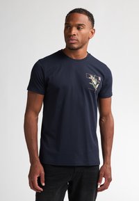 Marineblauw katoenen T-shirt met een bloemenafdruk op de linkerkant van de borst, ronde hals en korte mouwen. Eenvoudig en getailleerd ontwerp.