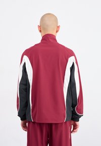 Sportjakke i bordeaux med sorte og hvide accenter. Har en høj krave, elastiske cuffs og en glat tekstur. Vist fra bagsiden.